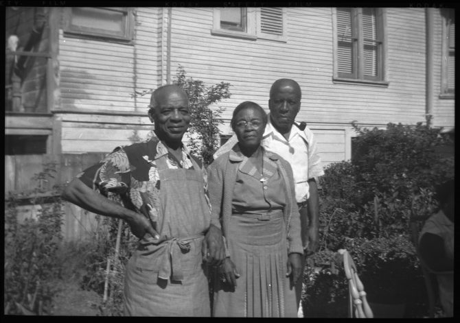 Three people stand bythe side of a house with shrubbery